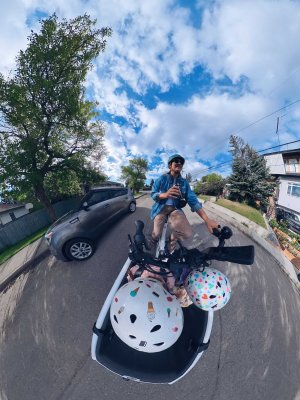 360 photo of two kids with colourful helmets in the front box of an urban arrow cargo bike while a woman pedals and drinks her coffee, smiling. The sky is blue with some white clouds and there are trees and homes in the background. 