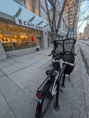 Urban Arrow cargo bike locked to bike rack on sidewalk by cafe, skyscrapers in background.