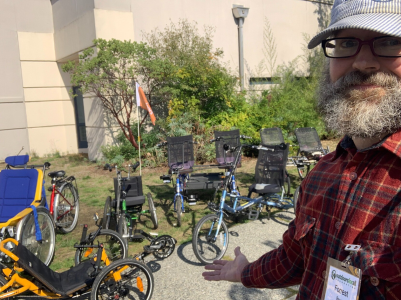 A guy with a beard and hat standing in front of a large variety of adaptive cycles.