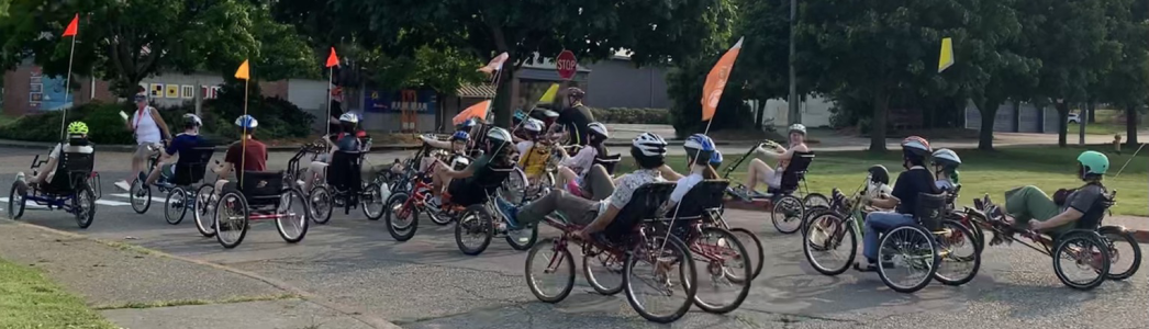 20+ people on various adaptive handcycles, tricycles, tandems, side-by-side bikes, or therapy trikes (many with flags for visibility).